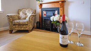 A living room with a floral armchair, a wooden table with a wine bottle and glasses, a fireplace, and a television mounted on the wall. - Gorgeous, fully refurbished centrally located cosy cottage (Hawick)
