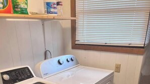 Un área de lavandería pequeña con lavadora y secadora de carga frontal, una ventana con persianas y un estante con productos de limpieza.