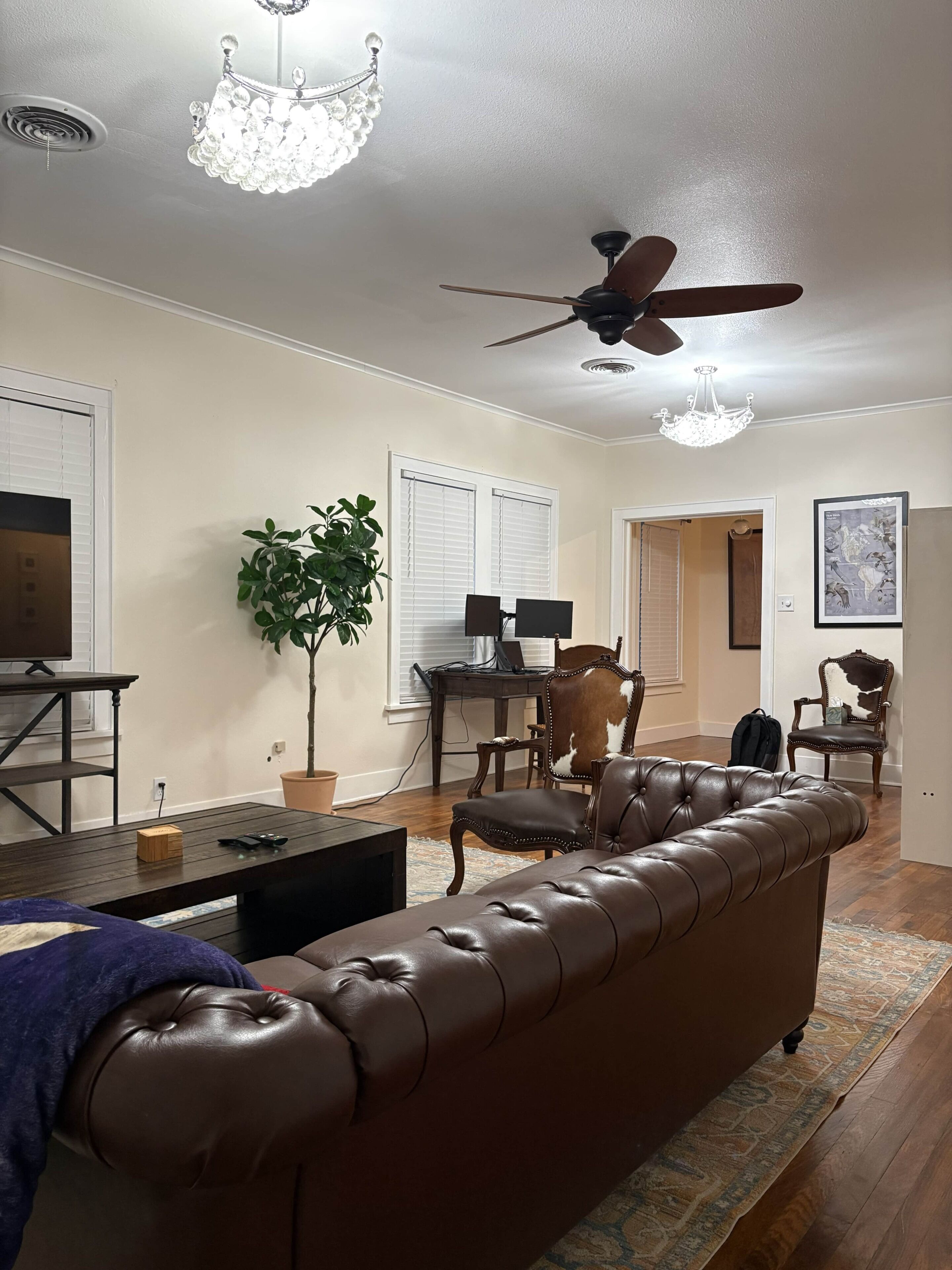A living room with a brown leather sofa, a dining table with chairs, a ceiling fan, and a chandelier.