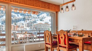 A dining area with a wooden table and chairs, a view of a snowy mountain town through large windows, and hanging light fixtures. - Nice apartment for 4 guests with WIFI, TV and balcony (Basse-nendaz)