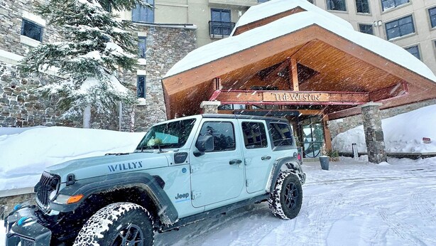 Un Jeep azul claro con neumáticos para todo terreno estacionado frente a un edificio con una gran entrada de madera y alrededores cubiertos de nieve.