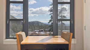 Un comedor con mesa y sillas de madera, ventanas amplias con vistas a un bosque y edificios.