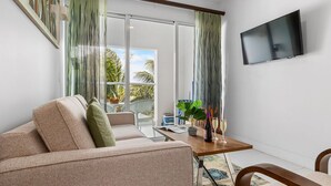 Een moderne woonkamer met een bank, een salontafel met een plant en een fles, een flatscreen televisie en een groot raam met gordijnen.