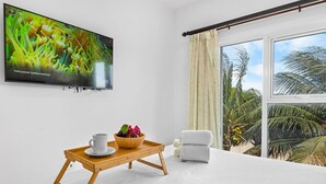 Een moderne kamer met een grote televisie, een houten dienblad met een kop en schaal, een witte stoel en uitzicht op palmbomen.