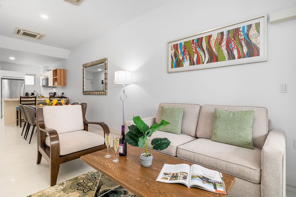 A modern living room with a sofa, coffee table, and chairs. There is a dining area in the background with a table and chairs.