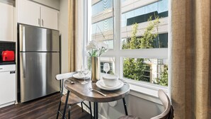A modern kitchen with a stainless steel refrigerator, a dining table with white plates and glasses, and a window with a view of greenery outside.