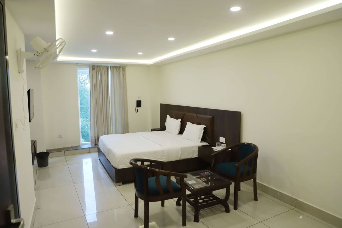 A hotel room with a large bed, a desk, two chairs, a small table, a ceiling fan, and a window with curtains.