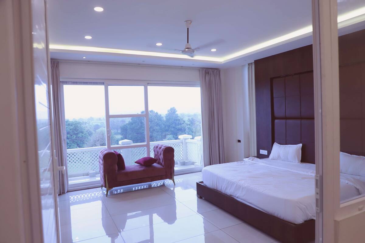 A modern bedroom with a large bed, a red armchair, and a balcony with a view of trees.