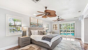 A bedroom with a large bed, two ceiling fans, a nightstand, a lamp, and a view of a swimming pool through glass doors. - Canal Access: Boat Lift, Heated Pool & Dock (Cocoa Beach)