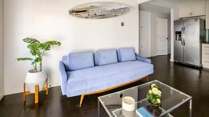 En moderne stue med en blå sofa, et glasbord, en potteplante og et køkken i baggrunden.