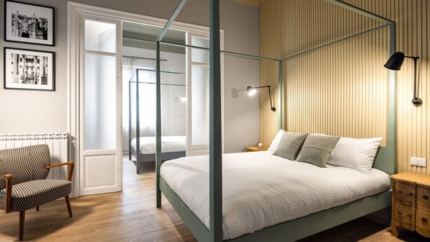 A bedroom with a four-poster bed, a wooden nightstand, a chair, and framed pictures on the wall. - Constance Luxury Apartment by DomuSicily (Palermo)