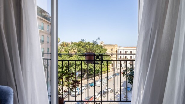 A balcony with a view of a busy street, a building with a balcony, and a clear sky. - Palazzo Fatta Apartment by DomuSicily (Palermo)