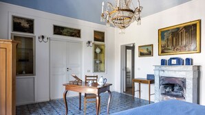 A room with a bed, a wooden table with chairs, a chandelier, a fireplace, and paintings on the walls. - Palazzo Fatta Apartment by DomuSicily (Palermo)