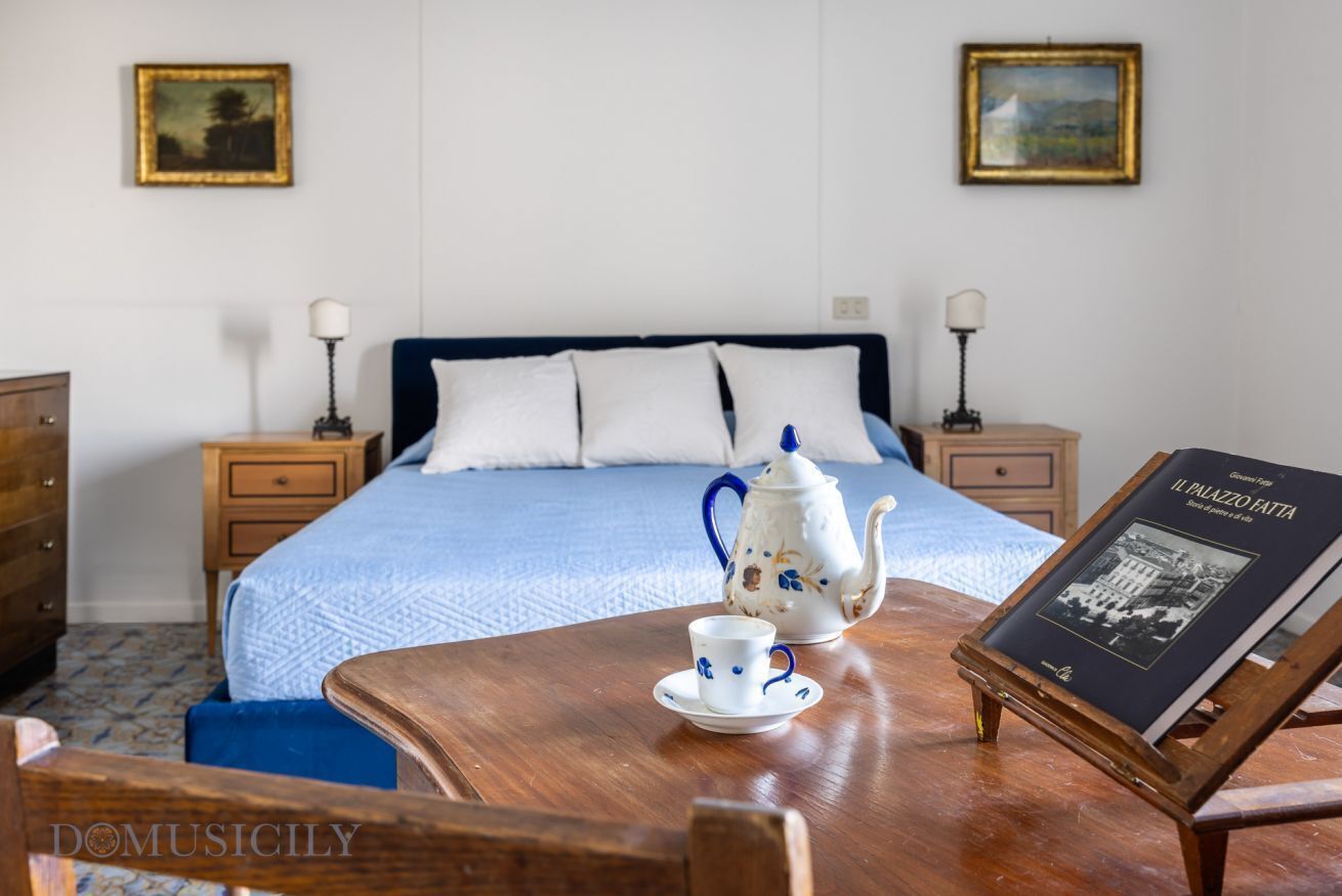 Ein ordentlich eingerichtetes Schlafzimmer mit einem Bett, Nachttischen, einem Holztisch mit Teekanne und Tasse sowie zwei gerahmten Bildern an der Wand.