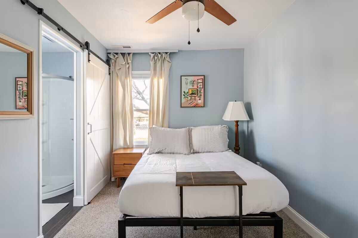 Un dormitorio con cama, ventilador de techo, mesita de noche, lámpara, ventana con cortinas y puerta que da a un baño.