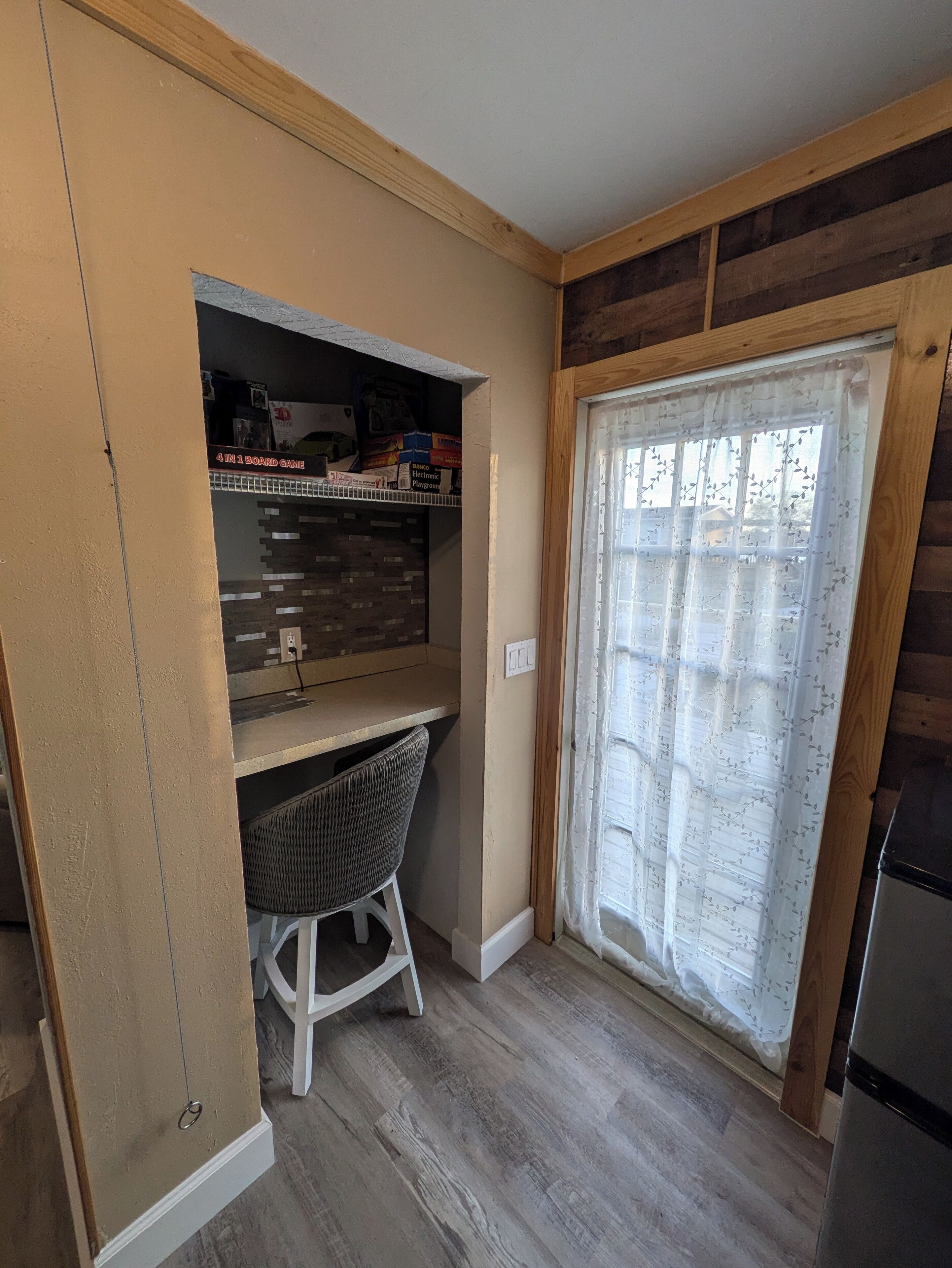 A compact kitchen with a built-in desk, a wicker chair, and a window with a floral curtain.