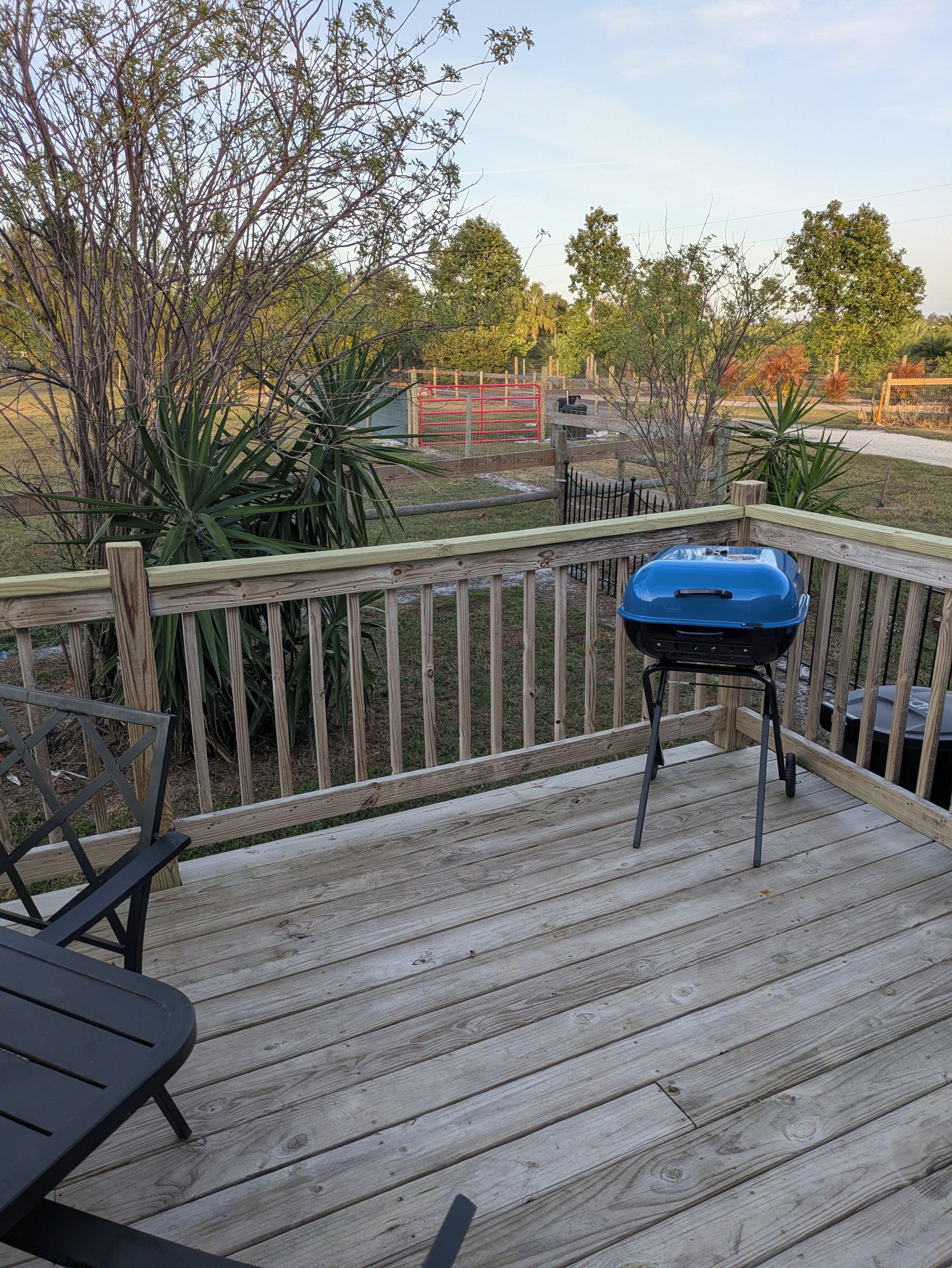 Une terrasse en bois équipée d’un barbecue, d’un banc et offrant une vue sur une courClose par une clôture, avec des arbres et un portail rouge.