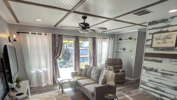 A modern living room with a sofa, a coffee table, and a TV. There is a ceiling fan, a window with curtains, and a wall with a framed picture.