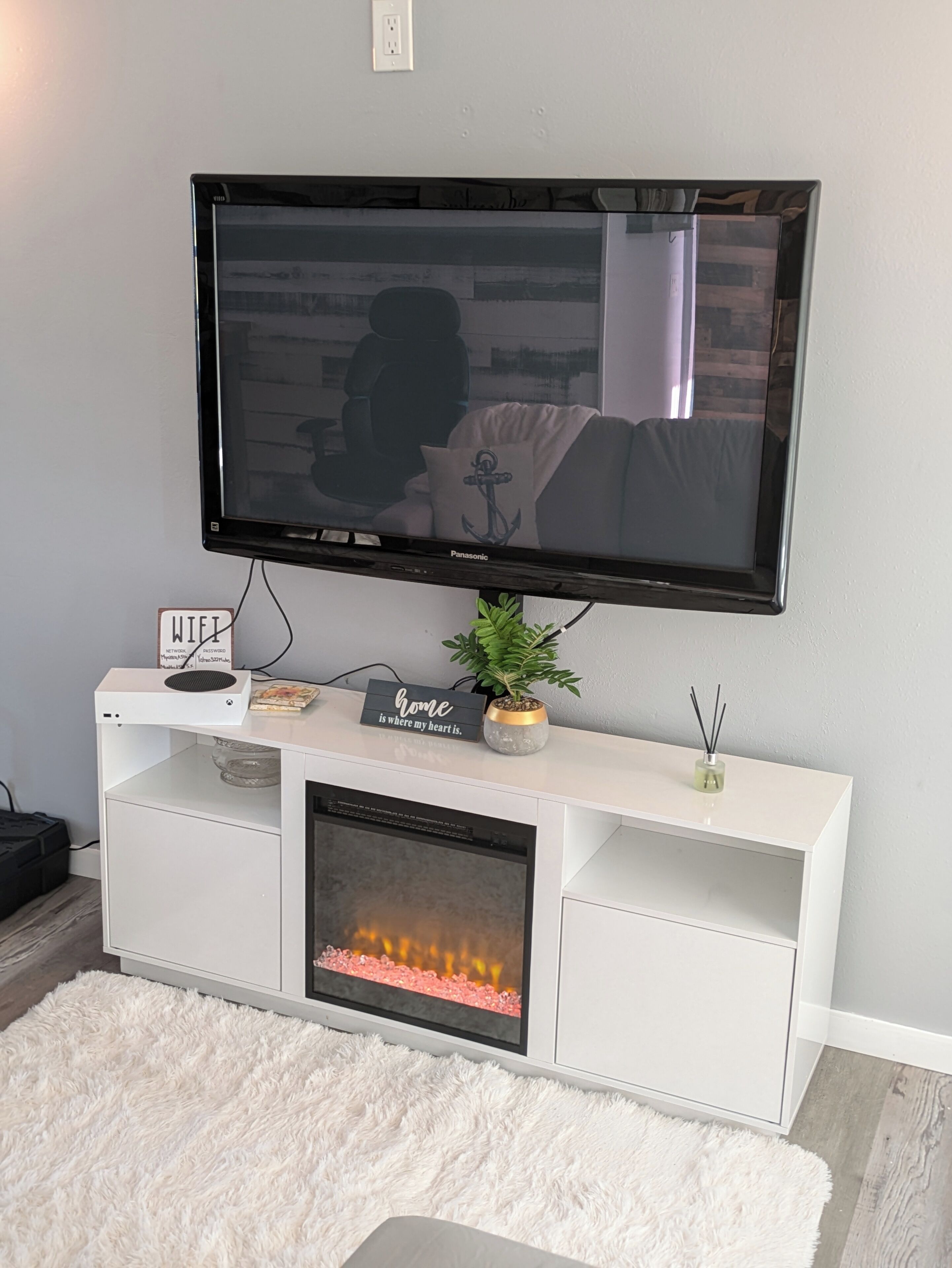 A modern living room with a flat-screen TV mounted on a white entertainment unit, a small potted plant, a sign reading "Home is where my heart is," and a fireplace below the unit.