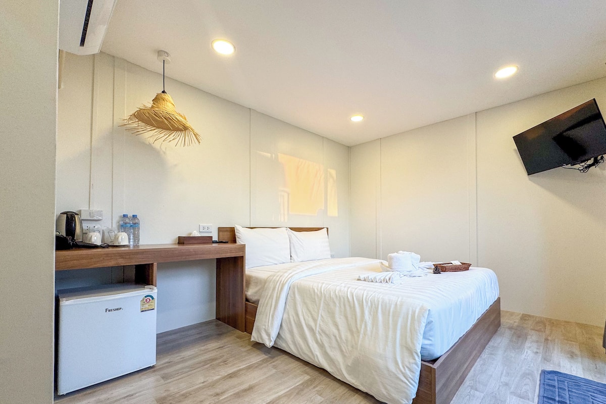 Una habitación de hotel con cama, una kitchenette pequeña, un televisor montado en la pared y una lámpara en el techo.