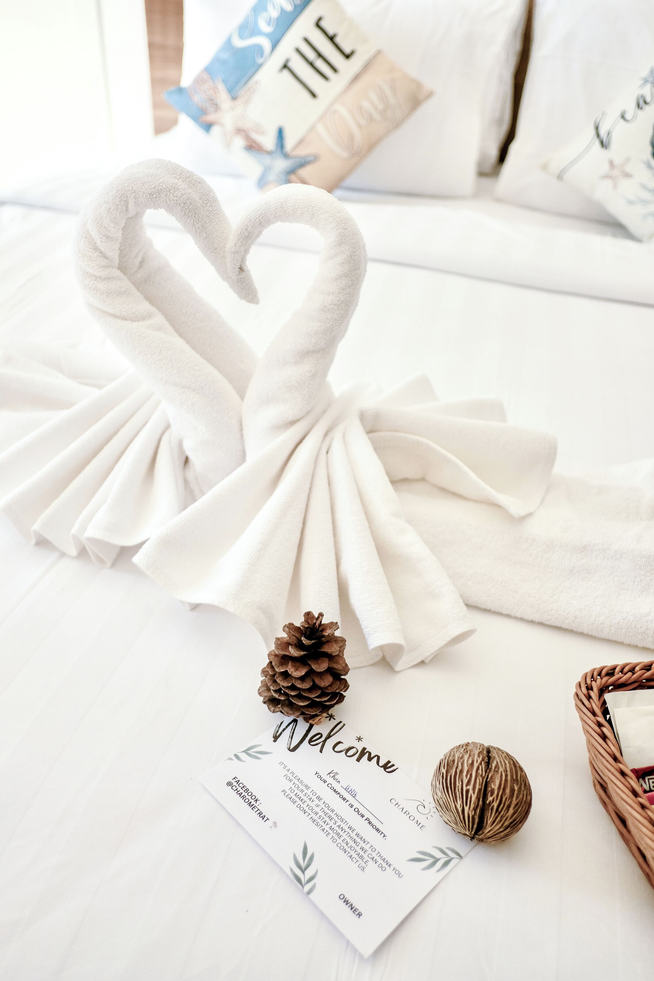 Un cœur en forme de serviette blanche est posé sur le lit, accompagné d’une carte de bienvenue, d’une pomme de pin et d’une petite boule décorative.
