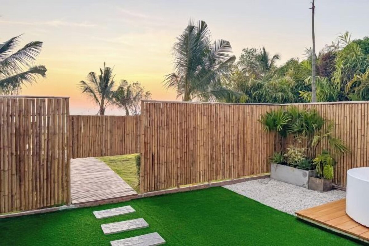 Un patio trasero con cerco de madera, césped artificial, piedras para pisar y una jardinera con plantas.