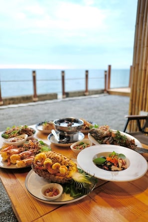 Mesa dispuesta con diversos platos y una olla caliente, con vista a una playa y una valla de madera.