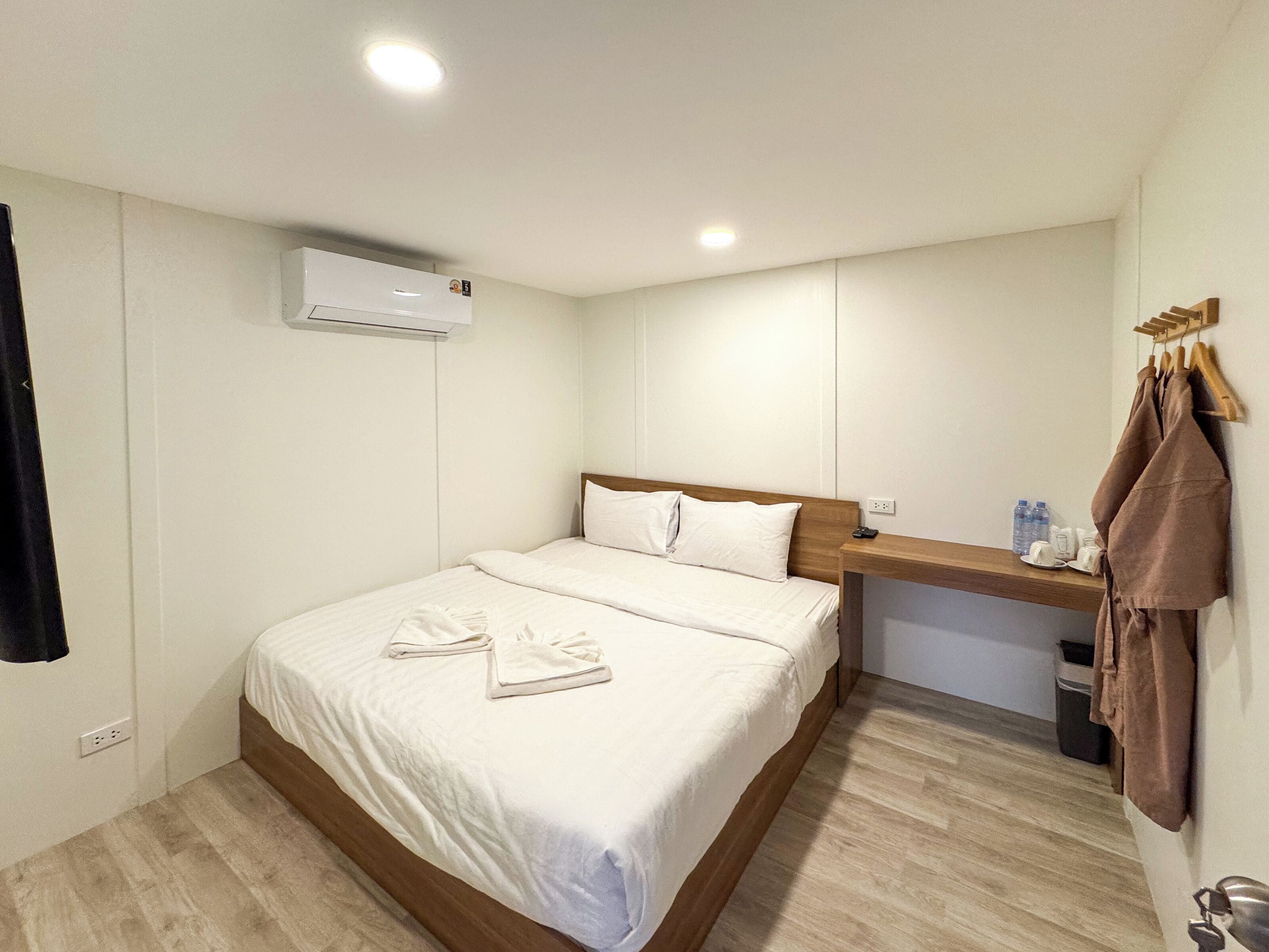 A hotel room with a bed, a wooden headboard, a bedside table, a wall-mounted air conditioner, and a towel rack.