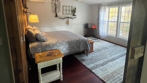 A bedroom with a bed, a nightstand, a ceiling fan, a window with curtains, and a wooden floor. - By The Rivers Edge....remodeled cozy cottage (Geneva)