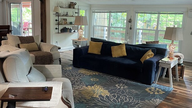 A spacious living room with a dark blue sofa, white armchair, and a patterned rug. There are two ceiling fans, a wooden floor, and a large window providing natural light. - By The Rivers Edge....remodeled cozy cottage (Geneva)