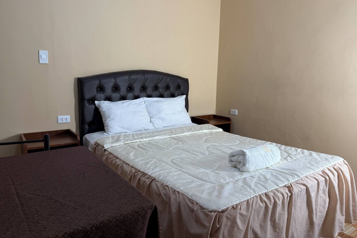 Ein ordentlich bezogenes Bett mit weißer Decke und Kissen, ein dunkles Kopfbrett und ein Nachttisch mit einer Lampe.