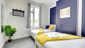 A hotel room with two beds, a window with curtains, a plant, and a painting on the wall.