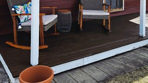 Un porche abrité avec deux fauteuils en osier et une petite table, une plante en pot, et une maison aux murs recouverts de bardage rouge et dotée d’une fenêtre.