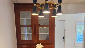 Un coin repas avec une table en bois foncé, un set de table à rayures et un vase contenant des fleurs séchées. On y trouve deux fauteuils rembourrés avec des détails dorés et un banc. Un lustre à plusieurs abat-jour noirs est suspendu au-dessus.