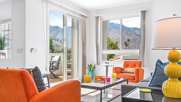 Ein modernes Wohnzimmer mit orangefarbenen Möbeln, einem Glastisch und Bergblick durch große Fenster.