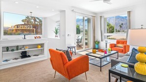 Ein modernes Wohnzimmer mit großem Flachbildfernseher, orange- und grauen Möbeln, einem Glastisch und Blick auf Palmen und Berge.