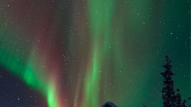 Une cabane aux murs en bois, entourée d’arbres couverts de neige, sous un ciel nocturne illuminé par les aurores boréales.