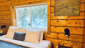Une chambre chaleureuse dans un chalet en bois, avec un grand lit, une fenêtre donnant sur une forêt et une lampe fixée au mur.