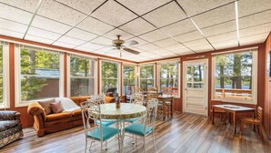 A sunlit room with a wooden floor, a round table with chairs, a sofa, and large windows. - Lakefront Lodge-Private Dock-Swimming-Game Room (Crivets)