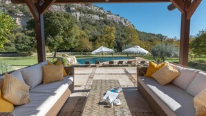 A covered outdoor seating area with wicker sofas and yellow pillows, a tiled table with a teapot, and umbrellas overlooking a pool and mountainous landscape.