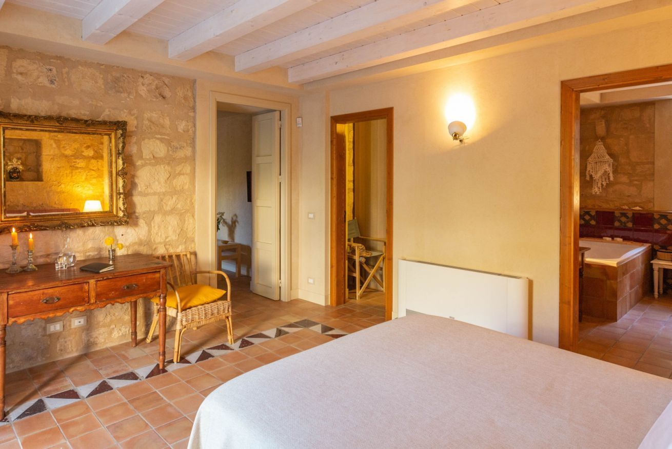 Una camera da letto con un letto, una cassettiera in legno, una sedia, uno specchio e una porta che dà su un'altra stanza.