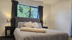Una cama bien hecha con cabecera gris y dos lámparas de noche, una ventana con cortinas y suelo cubierto de alfombra.