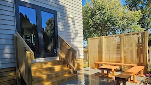 Una terraza de madera con un banco y una mesa, una escalera que lleva a una puerta, y una cerca de madera al fondo.