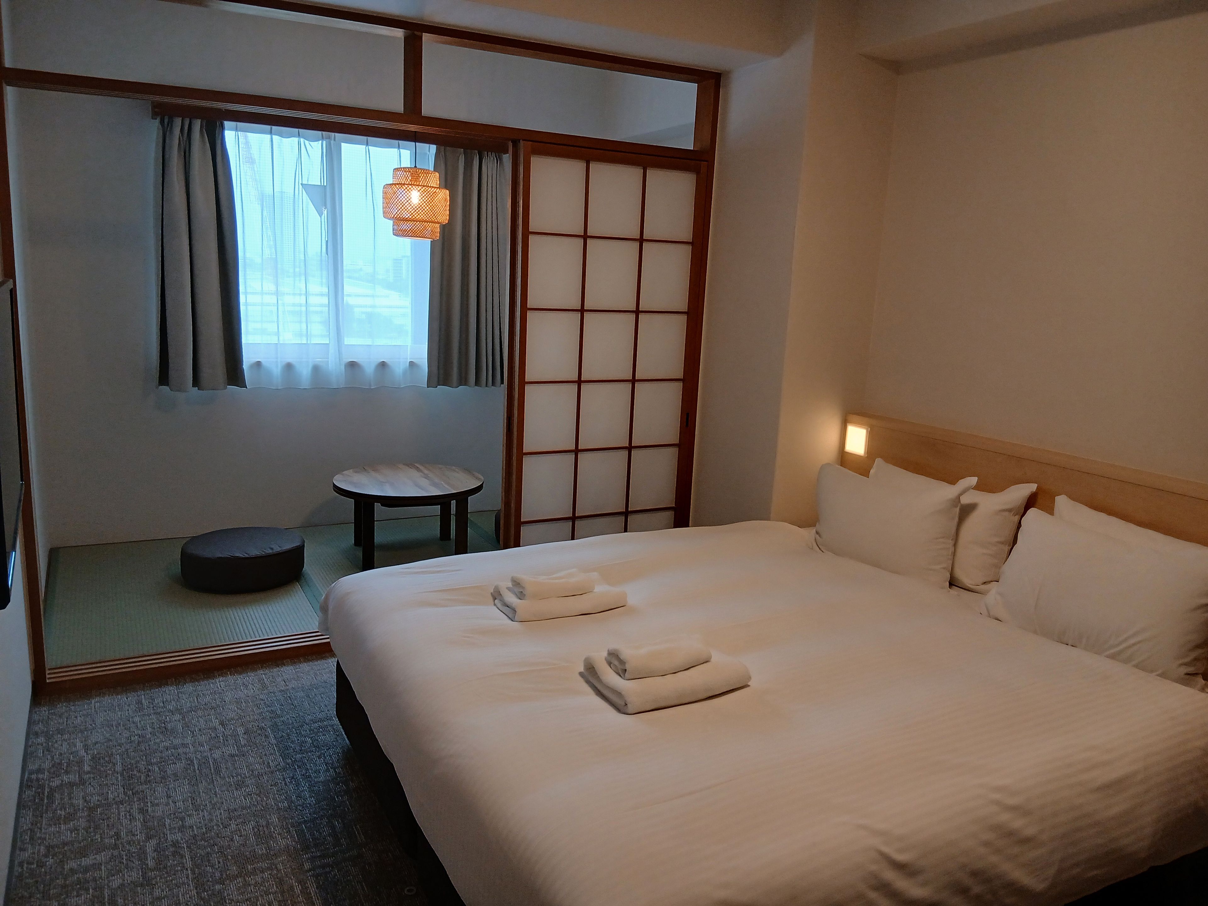 A hotel room with a bed, a small table, a chair, and a view through a shoji screen.