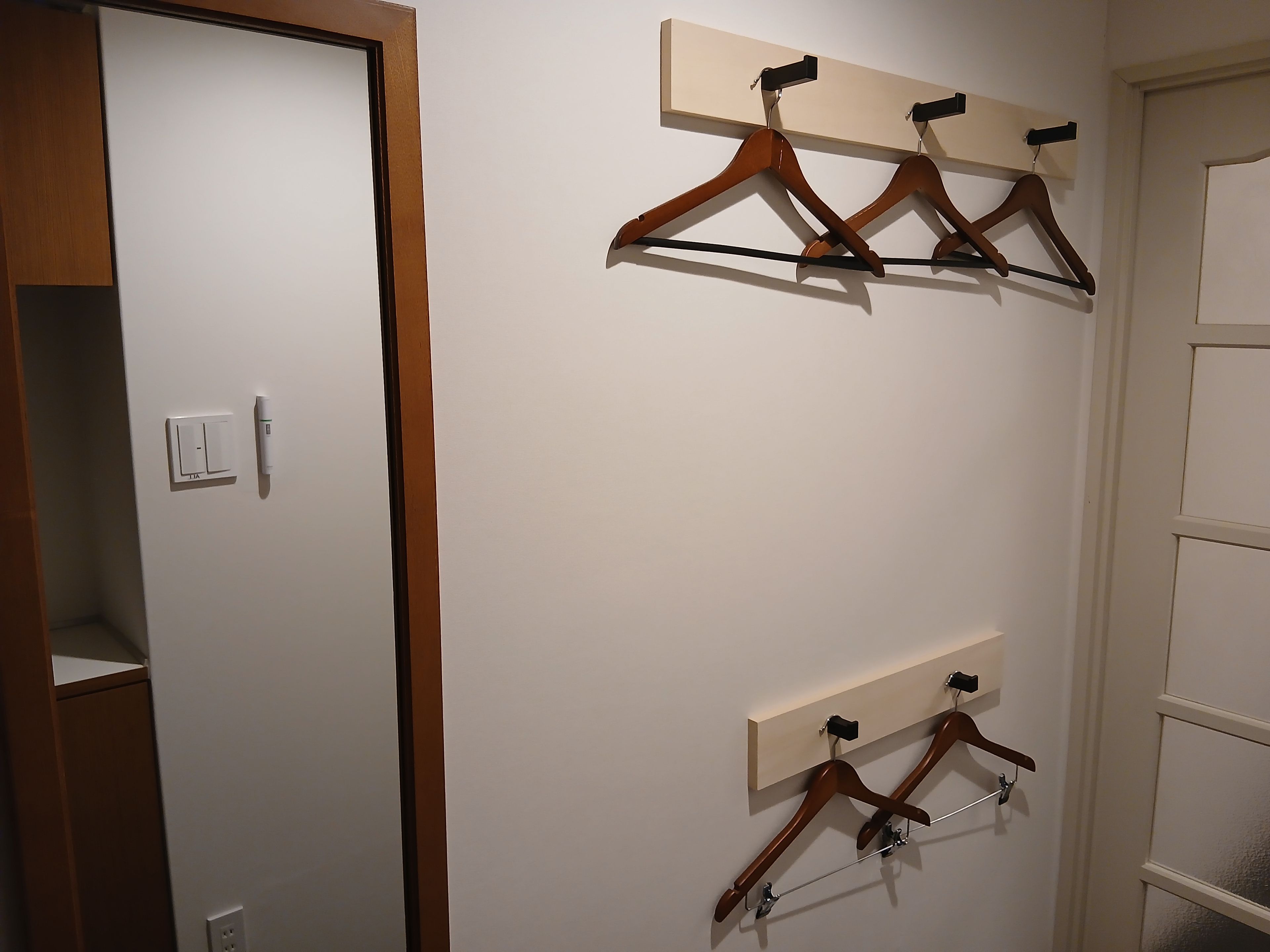 Una stanza con una porta bianca, un interruttore della luce e due grucce di legno fissate al muro.