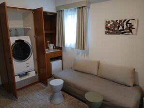 Ein kompakter Wohnraum mit Waschmaschine, einer kleinen Küchenzeile, einem Sofa und einem Einzelbett.