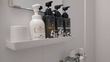 Ein Badezimmer mit einem weißen Regal, auf dem verschiedene Pflegeprodukte liegen, darunter eine Flasche Gesichts- und Handseife sowie mehrere Flaschen Shampoo und Duschgel.