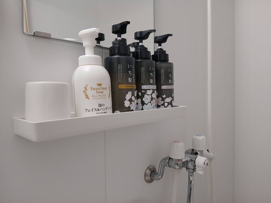 Ein Badezimmer mit einem weißen Regal, auf dem verschiedene Pflegeprodukte liegen, darunter eine Flasche Gesichts- und Handseife sowie mehrere Flaschen Shampoo und Duschgel.
