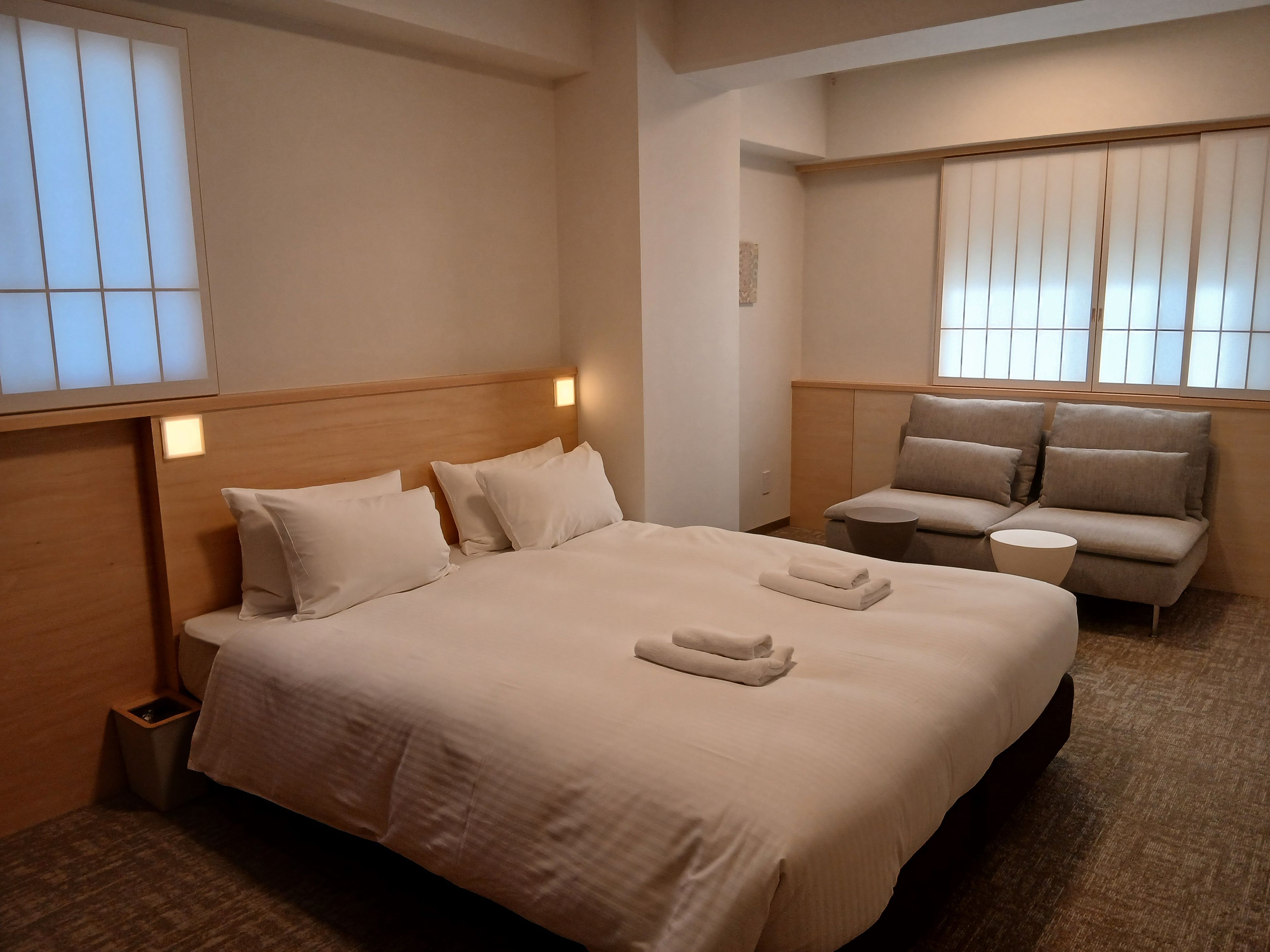 A hotel room with a large bed, a sofa, a small round table, and a window with blinds.