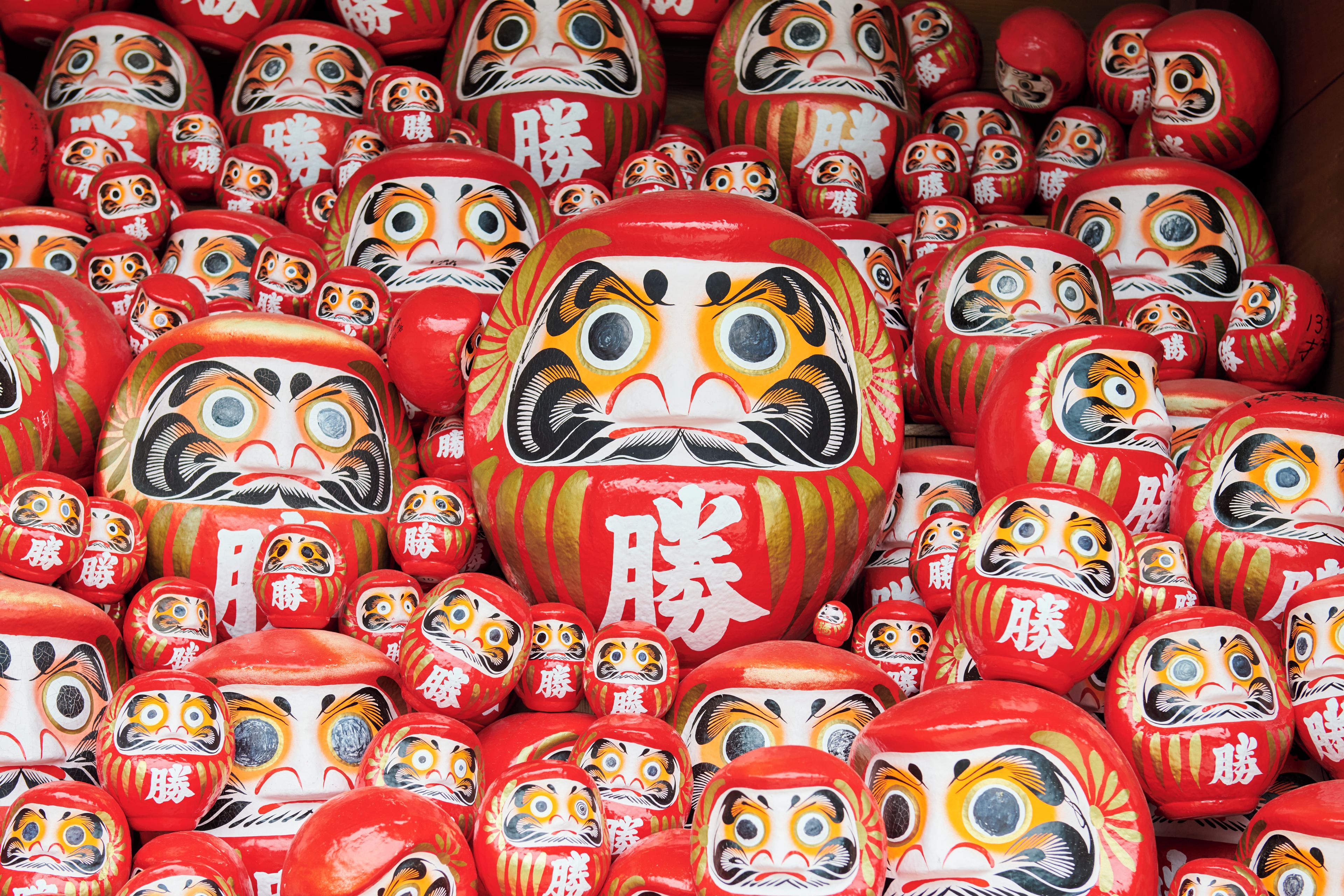 Une collection d’objets peints en rouge et en blanc, vraisemblablement des poupées traditionnelles, ornés de motifs complexes et dotés d’expressions faciales marquées.